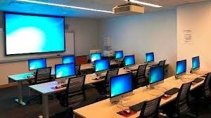 Computer lab with multiple desktop computers on desks facing a large projector screen in an office-like setting.